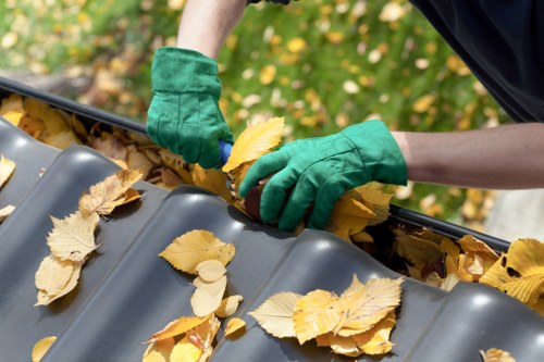 A close-up of a person wearing green work gloves collecting fallen yellow and brown leaves from a black plastic gutter system on a residential property. The gutter is partially filled with autumn foliage, with leaves scattered across its smooth, curved surface. The background features a blurred view of green grass and additional yellow fallen leaves, indicating an outdoor setting during the autumn season. This scene exemplifies routine maintenance involving clearing debris from guttering to prevent blockages, relevant to rubbish removal services offered by companies such as Commercial Waste Paddington, situated in the Paddington area within London postal districts. The image emphasizes the careful removal of organic waste material from property drainage systems, reflecting the practical aspects of waste management and domestic or commercial clearance activities throughout the UK, including Paddington.