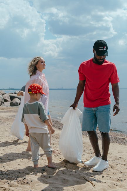 A man in a red t-shirt, denim shorts, and a black baseball cap stands on a sandy beach near the shoreline, holding a large white plastic bag, with a background of water and a partly cloudy sky. To his
