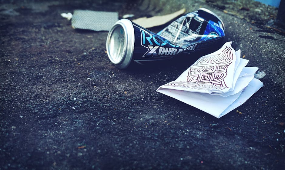 An orange plastic rubbish bag lying crumpled on a dark asphalt surface next to a grey stone curb in an outdoor setting. The bag appears to be partially torn or loose, with black printed text visible o
