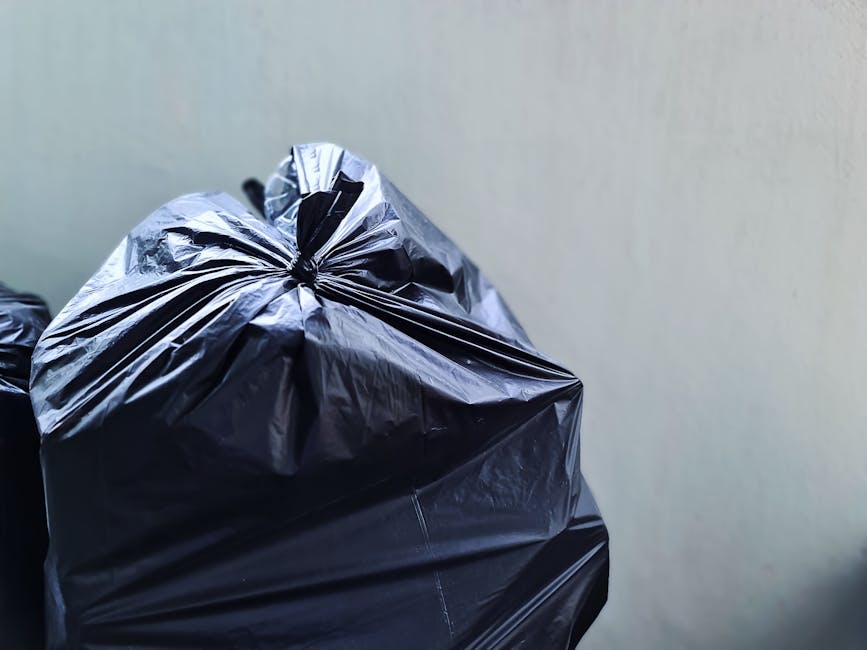 A middle-aged man with dark hair, wearing a black short-sleeved T-shirt and dark trousers, is seen disposing of rubbish into a metallic public waste bin on a paved pathway. The waste bin is cylindrica
